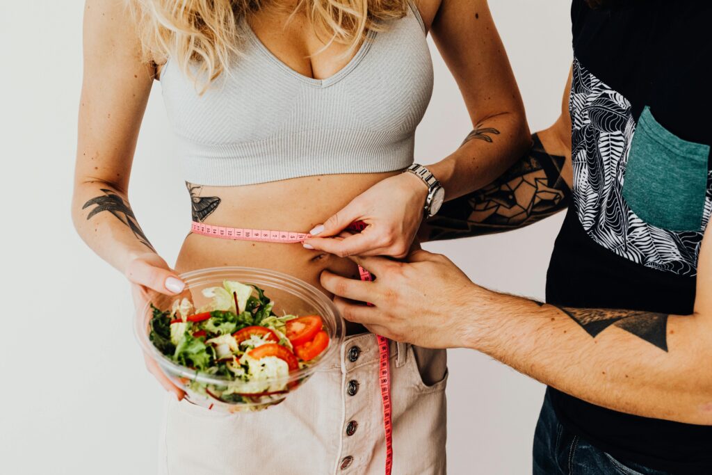 A couple measuring waistline with a tape and holding a fresh salad bowl, focusing on fitness and weight loss.